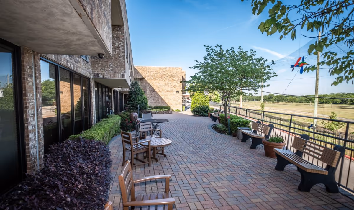 Brick-clad senior living outdoor terrace with patio tables, benches, potted plants and a railing overlooking open grounds.