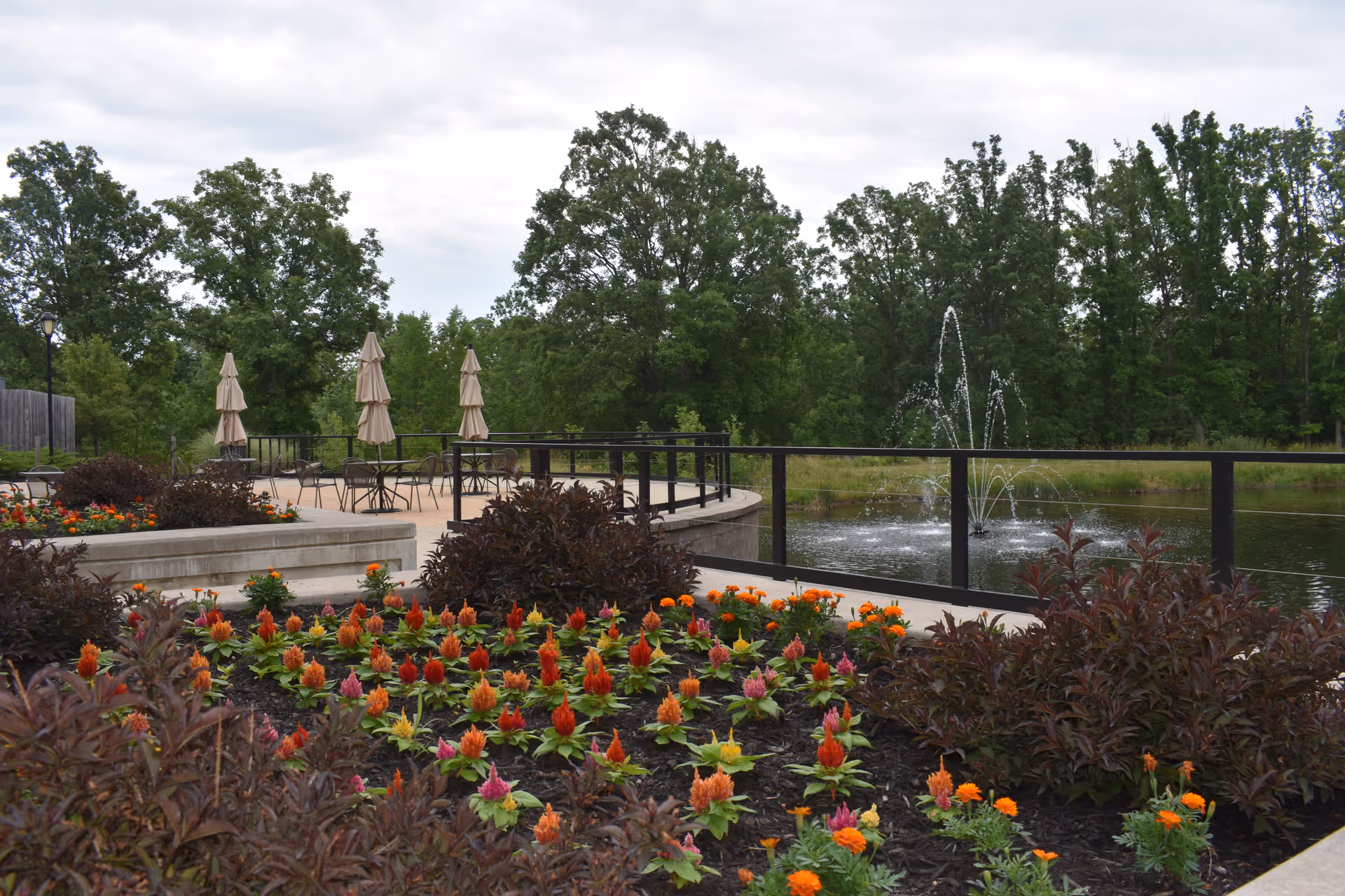 Landscaped outdoor patio with flower beds, tables and closed umbrellas beside a pond with a fountain.