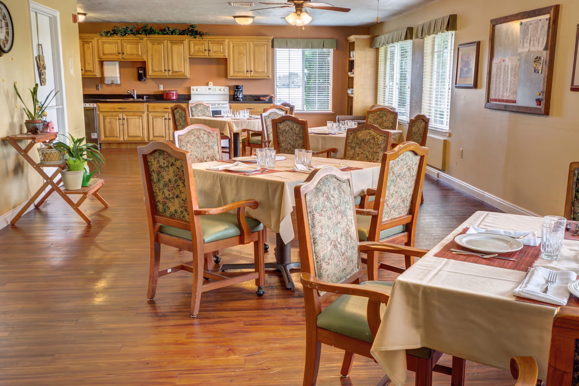 A dining room in a senior living facility with several tables covered in beige tablecloths, each set with plates, glasses, and napkins. The chairs have wooden frames with floral upholstered backs and green cushioned seats. The room has wooden flooring, large windows with blinds, and a kitchen area with wooden cabinets and appliances in the background. There are plants on a small wooden stand and framed pictures on the walls.
