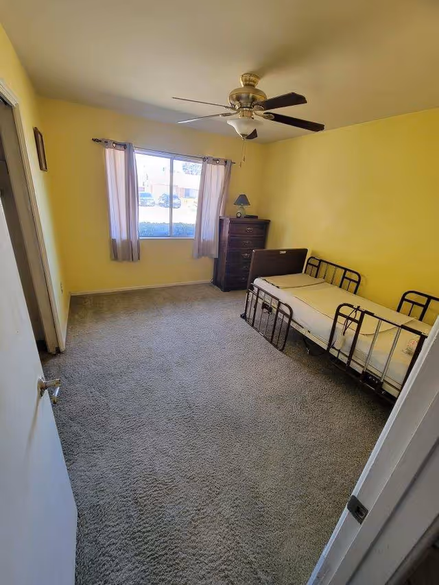 Small bedroom with yellow walls, a single bed with metal rails, a dresser by a window with curtains, and a ceiling fan.