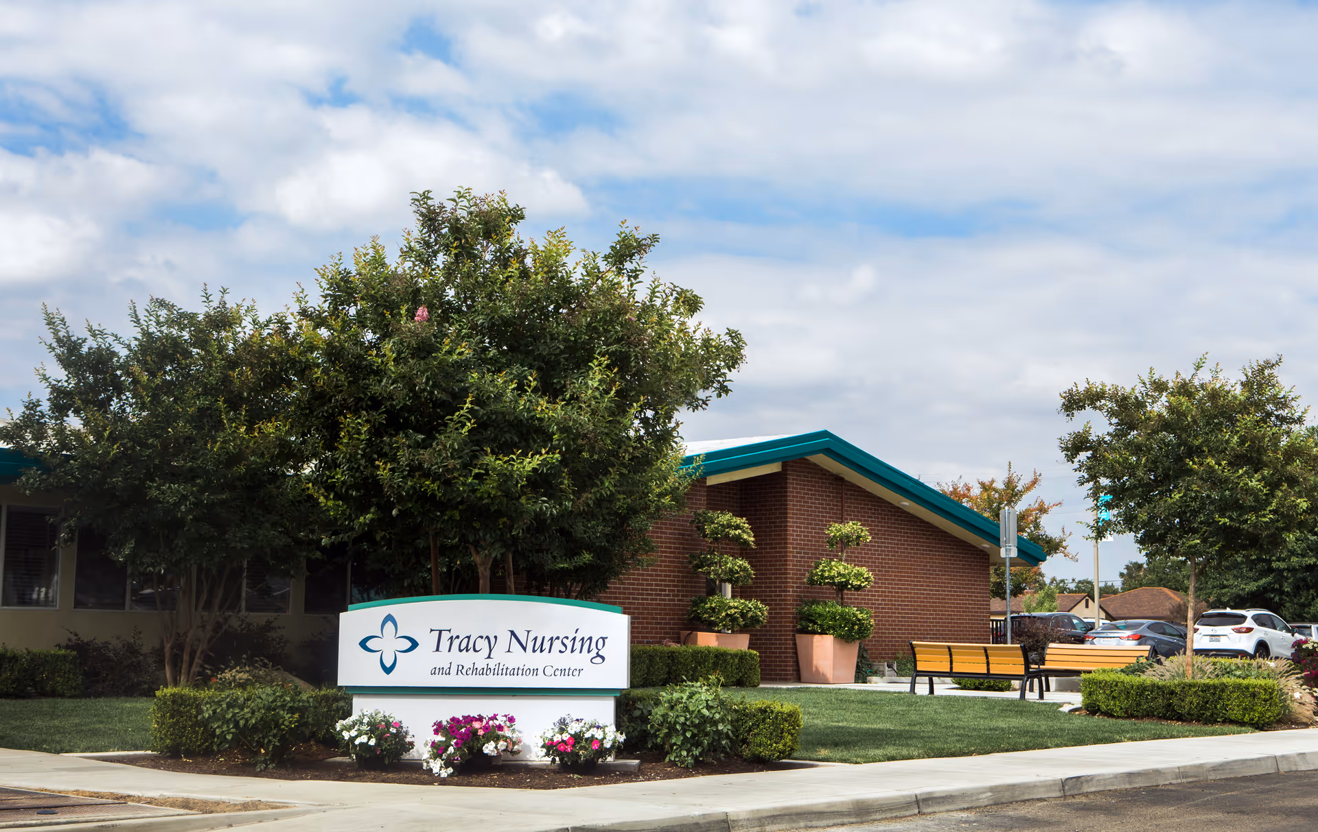 Front entrance of Tracy Nursing and Rehabilitation Center showing the sign, landscaped greenery, benches, and a brick building under a partly cloudy sky.