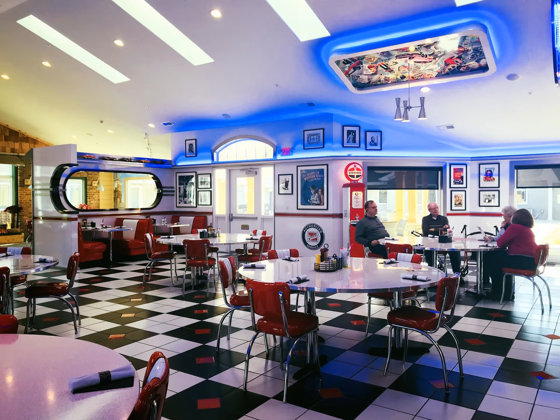 A retro-style dining room with black and white checkered floor tiles accented by red diamond shapes. The room features round white tables with red chairs and red booth seating along the wall. The walls are decorated with vintage posters and framed pictures, and there is a vintage gas pump near the back wall. Blue LED lighting highlights the ceiling, which has a colorful collage in a recessed area. Four elderly people are seated at a table near the window, engaged in conversation.