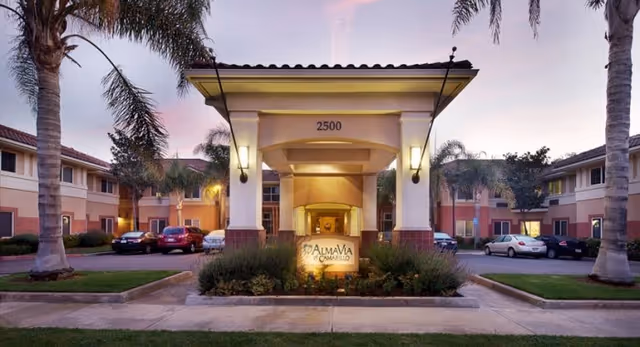 Front entrance of AlmaVia of Camarillo with archway, landscaping, and parked cars at dusk.