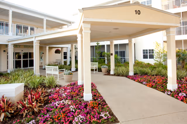Entrance area of a senior living facility with a covered drop-off zone marked with the number 10. The pathway is surrounded by vibrant flower beds and greenery, with white benches and potted plants near the building. The building has multiple windows and a balcony above the entrance.