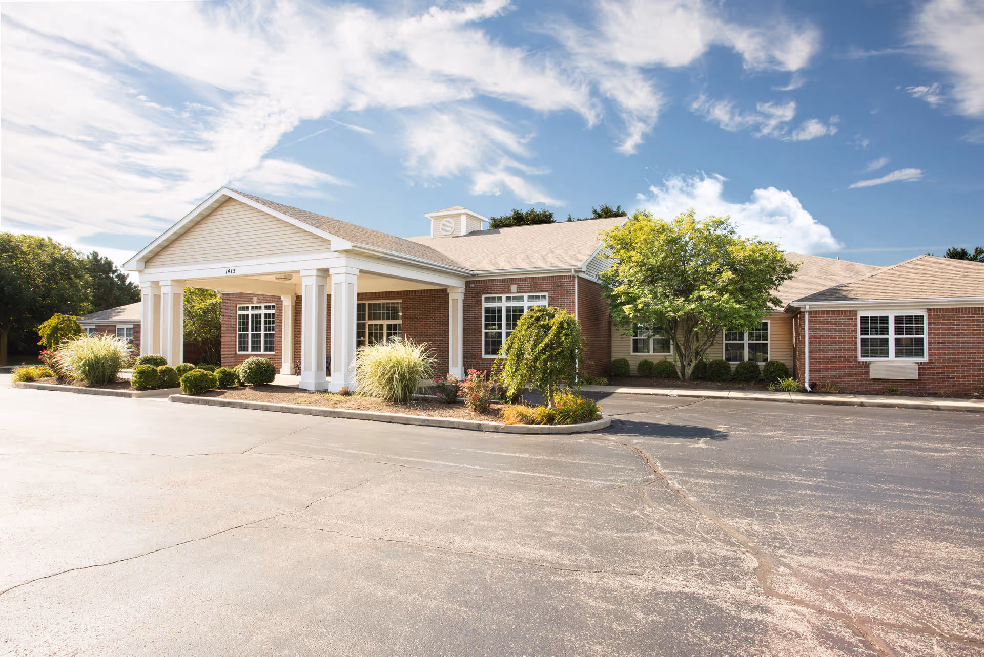 Single-story brick senior living facility with a columned covered entry, landscaped shrubs and a paved parking area under a blue sky.