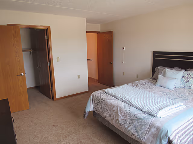 A simple bedroom with a made bed, open closet and doorway, beige walls and carpet.