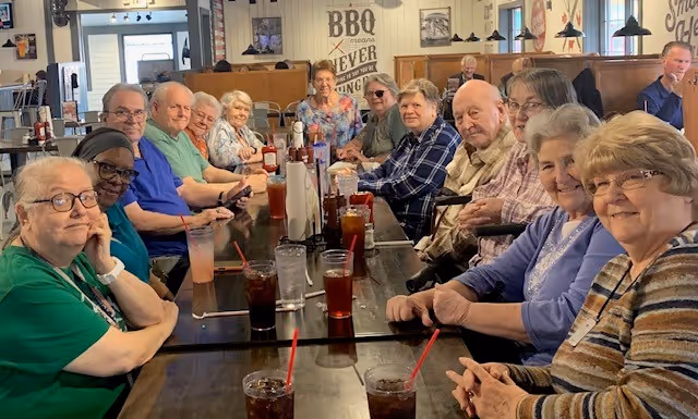 A group of elderly people sitting around a long dining table in a restaurant, smiling and enjoying drinks with red straws. The interior has wooden booths and walls decorated with framed pictures and a sign that reads 'BBQ means NEVER SAYING NO'.
