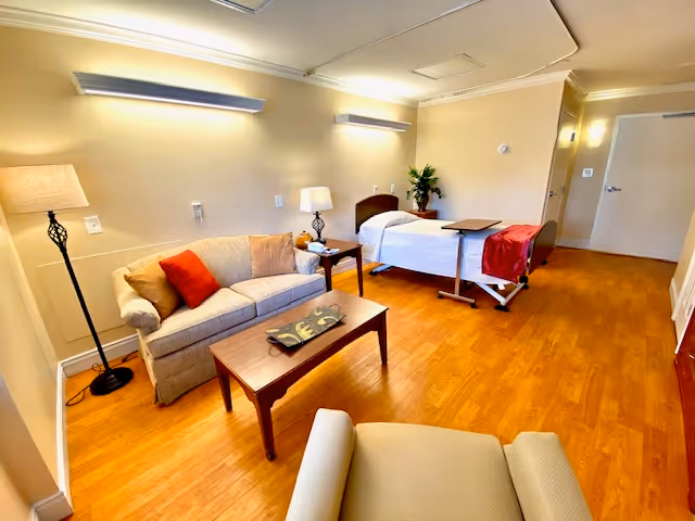 A well-lit room with a hospital-style bed covered with white bedding and a red blanket at the foot. Next to the bed is a small wooden side table with a lamp and some items on it. Across from the bed is a beige sofa with red and beige cushions, a wooden coffee table with a decorative tray, and a beige armchair in the foreground. The floor is wooden, and the walls are painted a light beige color with modern wall-mounted lights.