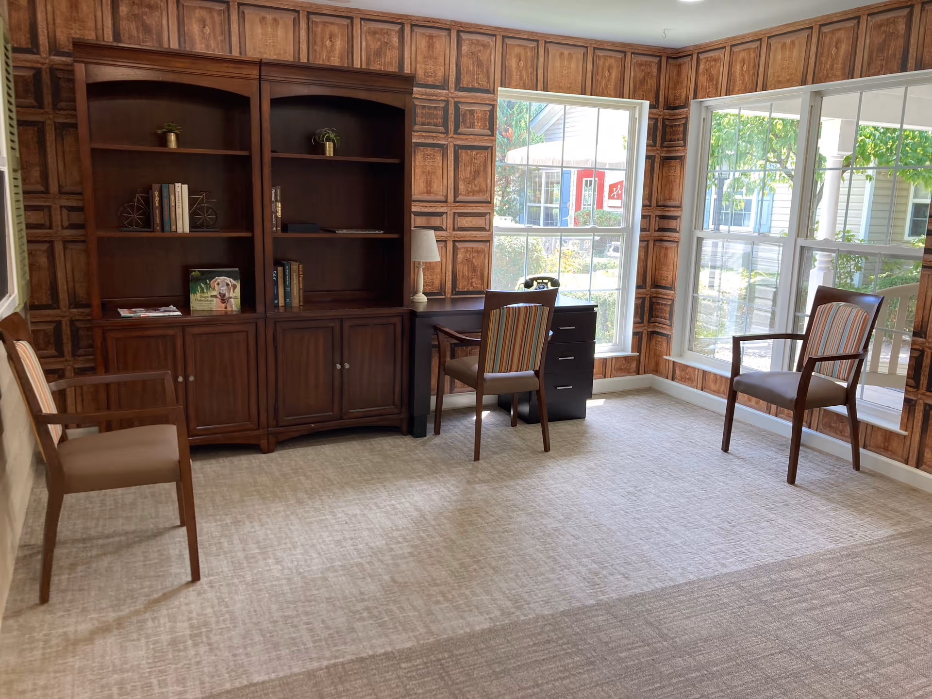 A bright room with large windows showing an outdoor view. Inside, there are three wooden chairs with striped cushions, a dark wooden bookcase with some books and small plants, and a small black desk with a lamp and a vintage telephone. The walls have a wood panel design, and the floor is carpeted.