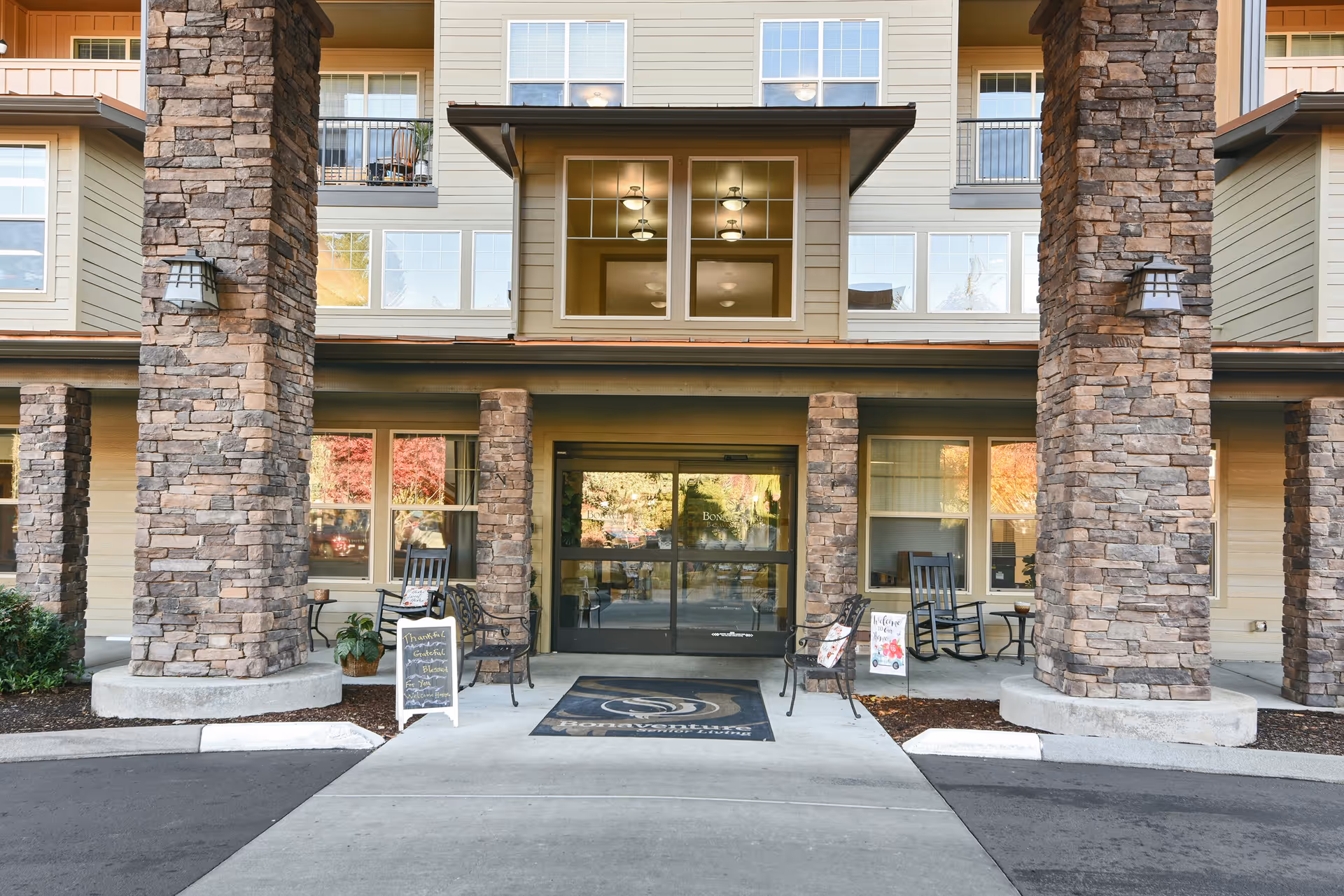 Entrance of a senior living facility with large stone pillars, glass double doors, rocking chairs, and welcome signs on either side of the walkway.