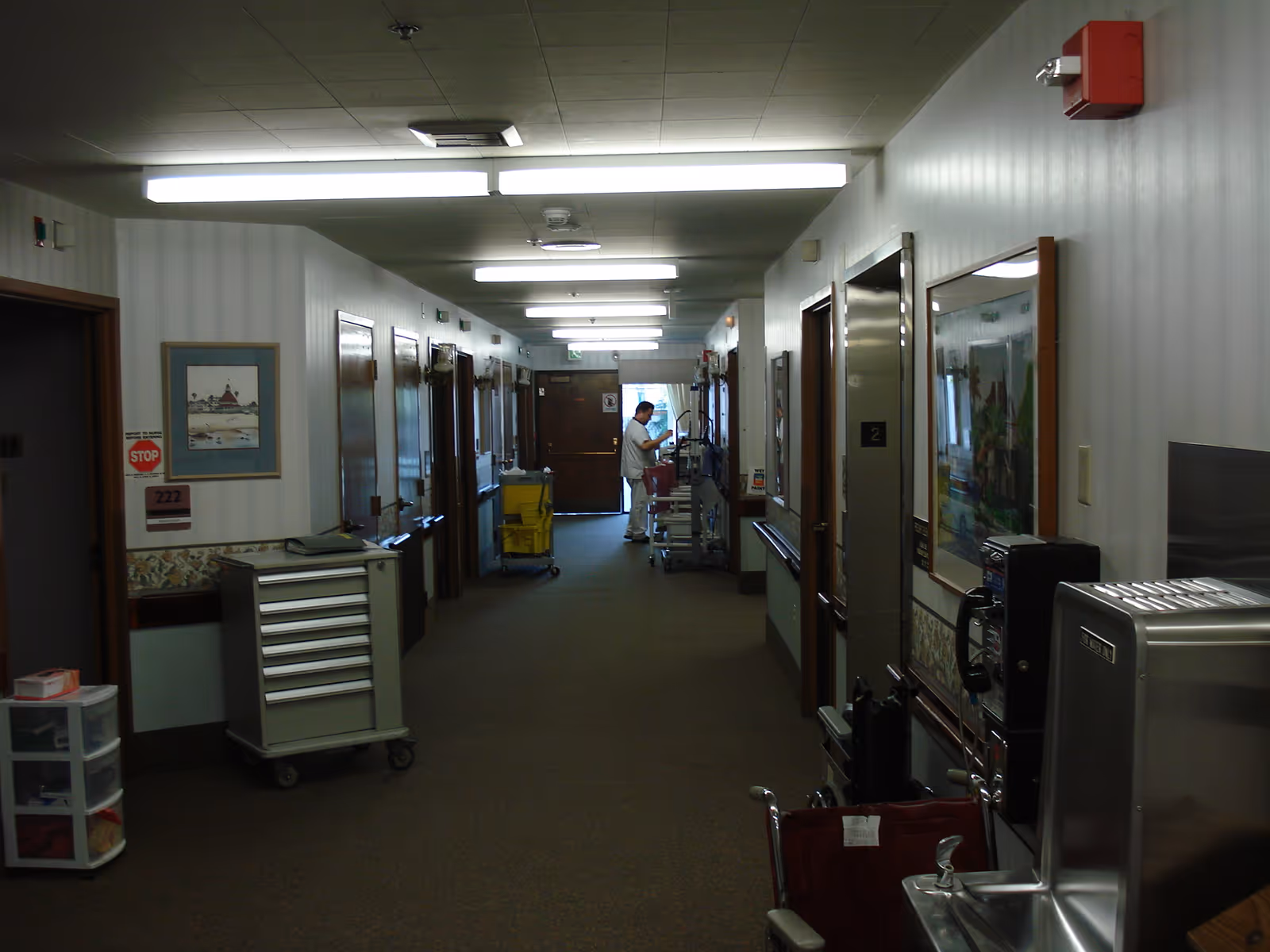 A long hallway in a skilled nursing and rehabilitation facility with closed doors on both sides, a person in white uniform working near the end of the hallway, medical carts, a wheelchair, and a water fountain visible. The walls have framed pictures and there are fluorescent lights on the ceiling.