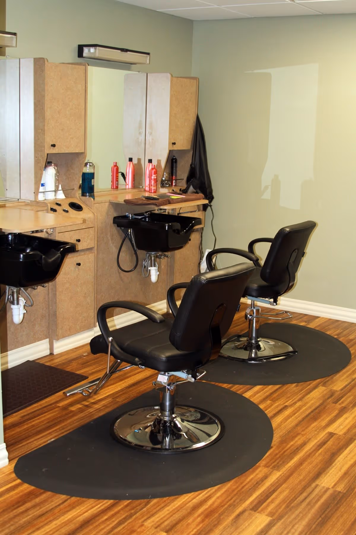Interior view of a salon area with two black salon chairs on black mats in front of two hair washing sinks and a counter with hair care products and a large mirror on the wall.