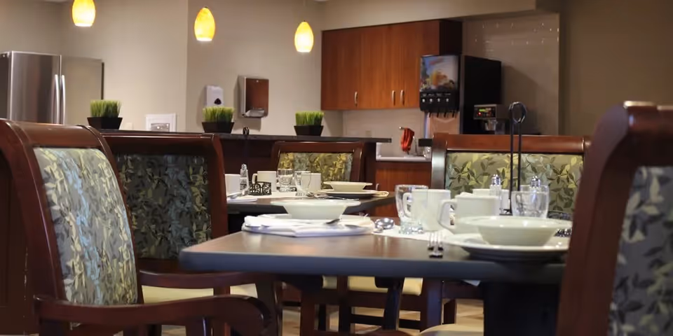 Dining area with tables set with white bowls, cups, glasses, and silverware. The chairs have wooden frames with green patterned upholstery. In the background, there are hanging pendant lights, a beverage dispenser, and wooden cabinets.