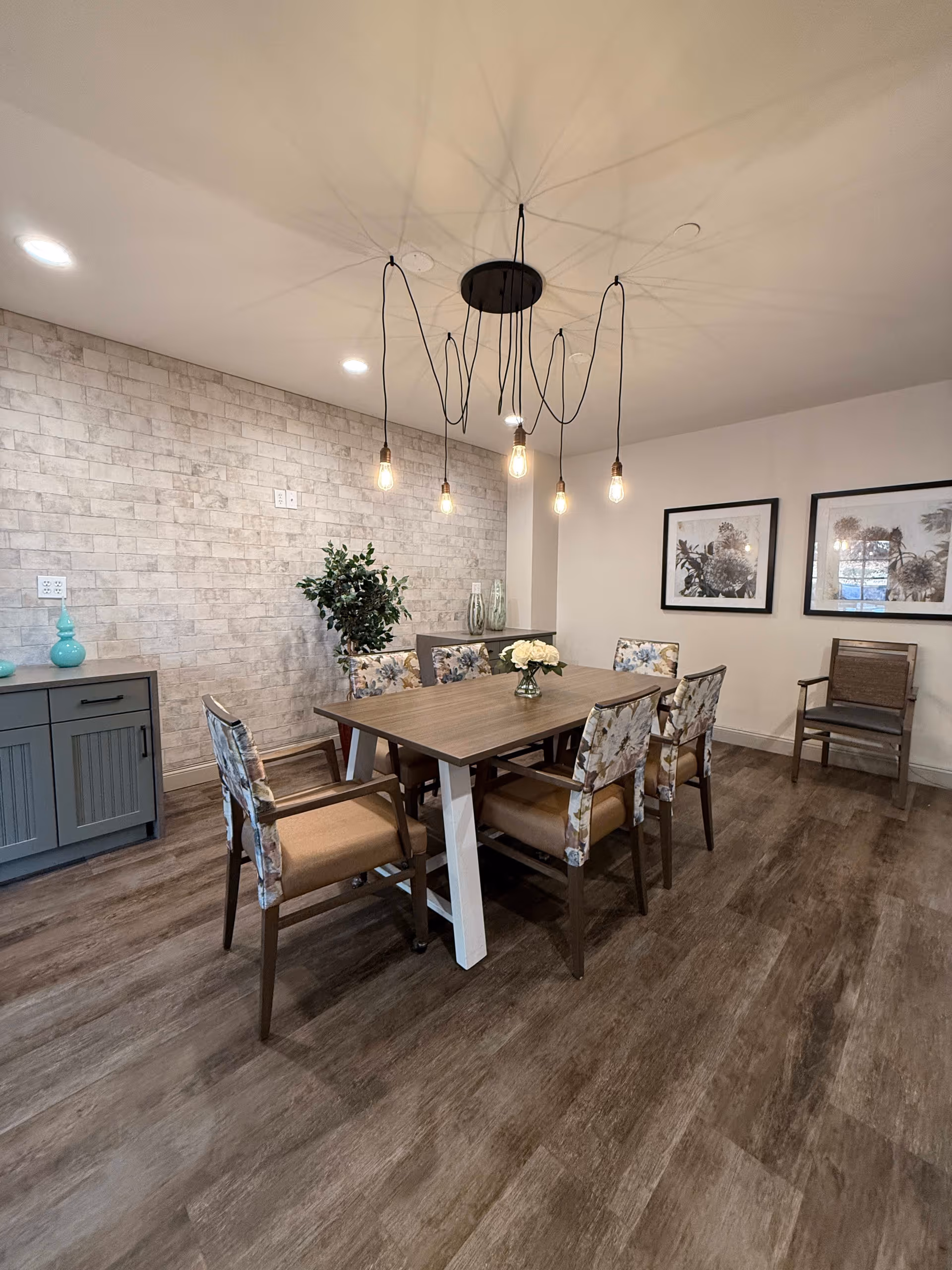 A modern dining room with a rectangular wooden table surrounded by six chairs with floral upholstery. Above the table hangs a unique chandelier with exposed light bulbs and black cords. The room features wood flooring, a light brick accent wall, two framed botanical prints on a plain wall, and a sideboard with decorative vases and a potted plant.
