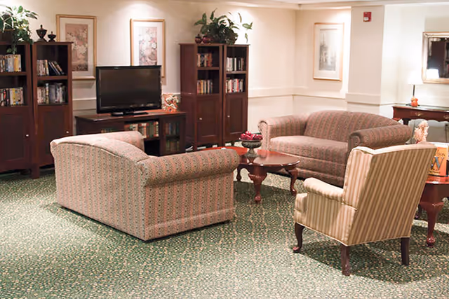 A cozy living room area with two patterned sofas and a striped armchair arranged around a wooden coffee table. There are bookshelves with books and decorative items, a TV on a stand, framed artwork on the walls, and a green patterned carpet covering the floor.
