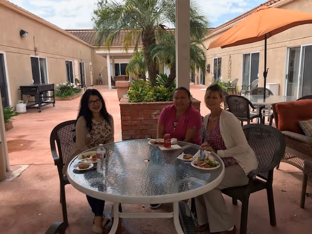 Three women sitting around a round glass table outdoors in a courtyard area with plates of food and drinks. The courtyard is surrounded by a single-story building with beige walls and windows. There are plants and palm trees in the center of the courtyard, and an orange umbrella providing shade over part of the seating area.