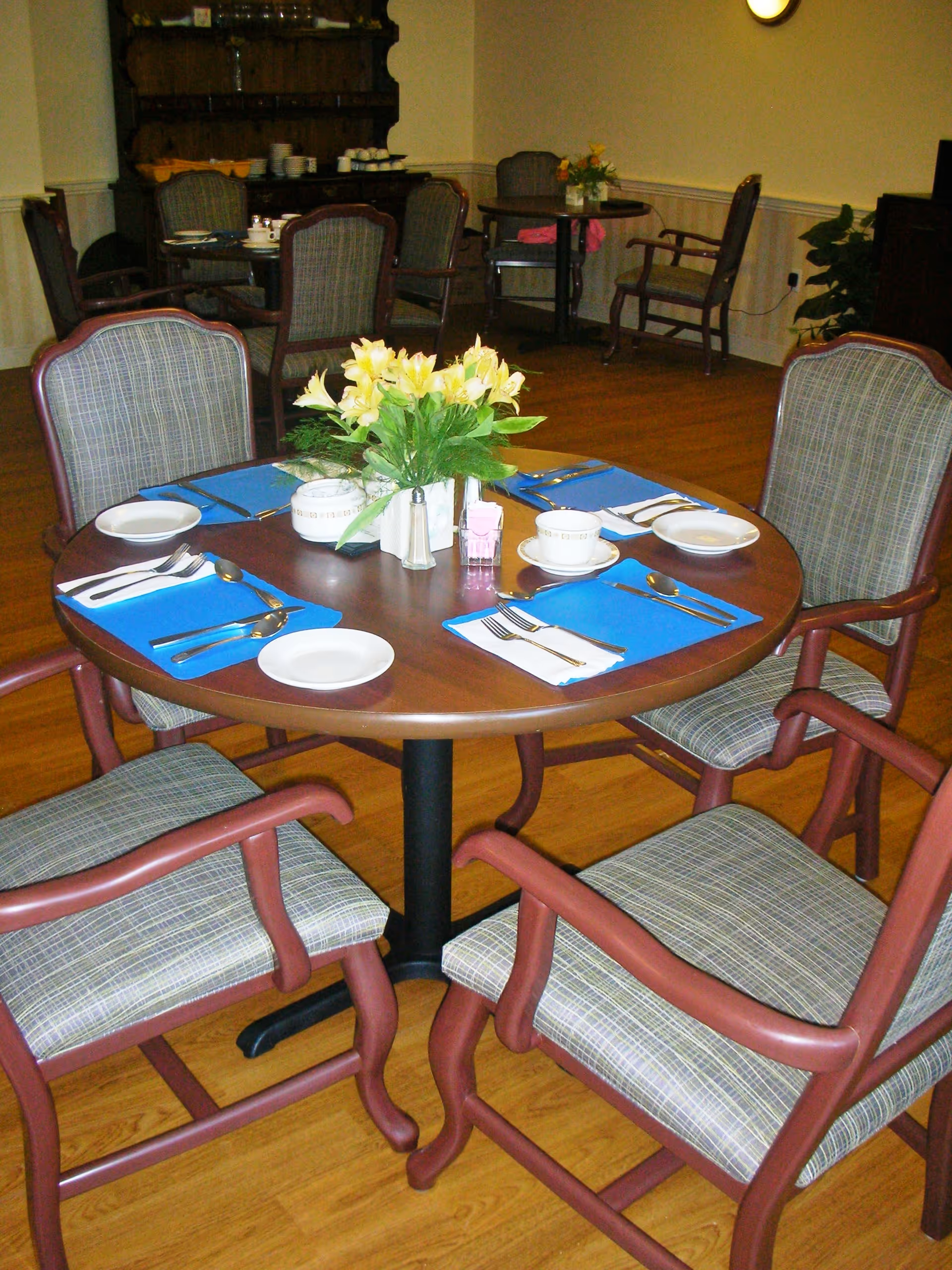 A round wooden dining table set for four with blue placemats, white plates, cups, and silverware. A small flower arrangement is in the center of the table. The room has wooden flooring and several other tables and chairs in the background, indicating a dining area.
