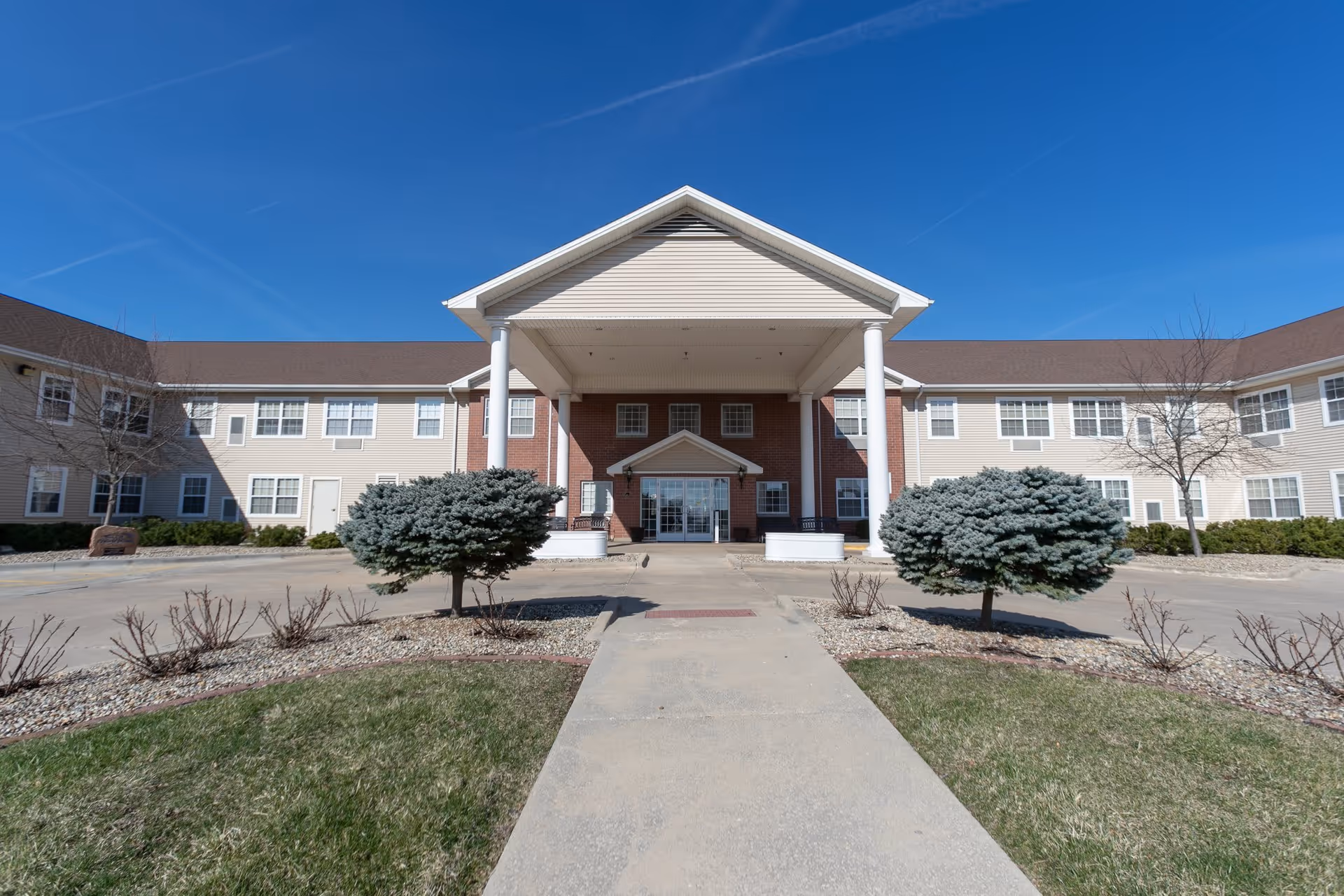 Front exterior view of Prairie Winds of Urbana facility showing a two-story building with beige siding and red brick accents, a covered entrance supported by white columns, landscaped bushes, and a clear blue sky.