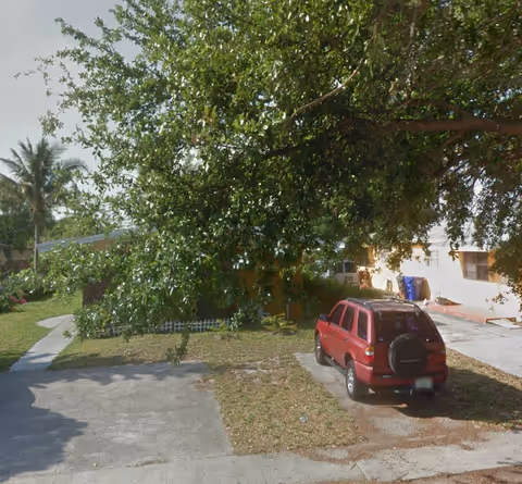 Front yard and driveway of a single-story building with a large tree and a red SUV parked near the entrance.