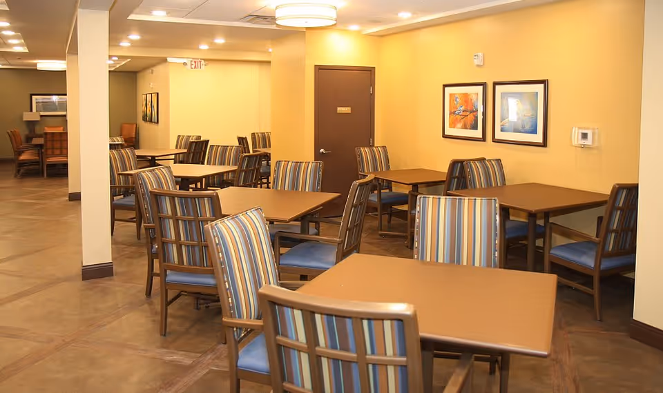 A dining area in an assisted living facility with multiple square wooden tables and chairs with striped upholstery. The walls are painted yellow and decorated with framed artwork. There is a door labeled 'Storage' and an exit sign above another door in the background. The floor is a polished brown surface.