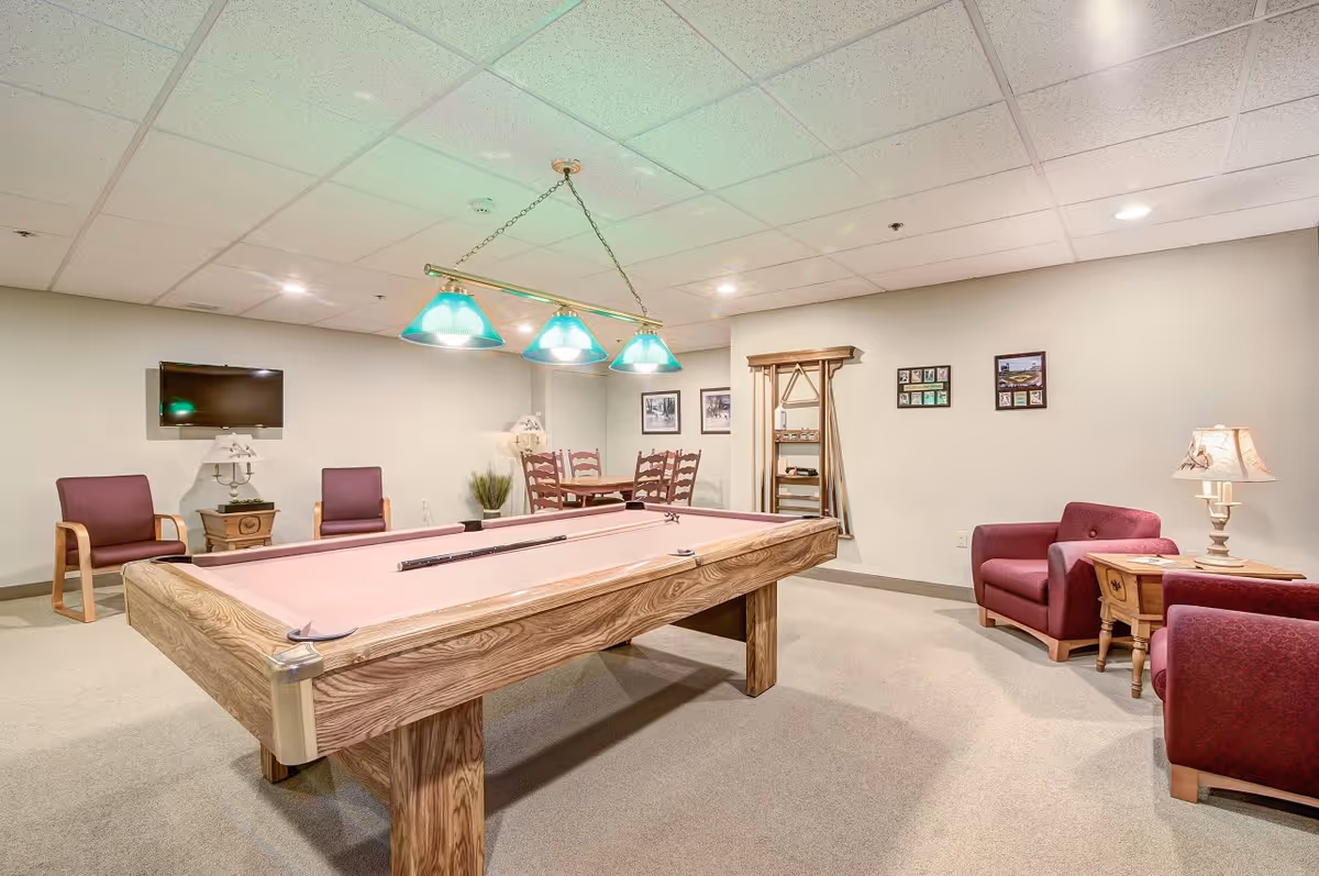 A recreational room with a pool table in the center, surrounded by several chairs and a small table with a lamp. There is a wall-mounted TV, framed pictures on the walls, and a rack holding pool cues. The room has a carpeted floor and a drop ceiling with recessed lighting.