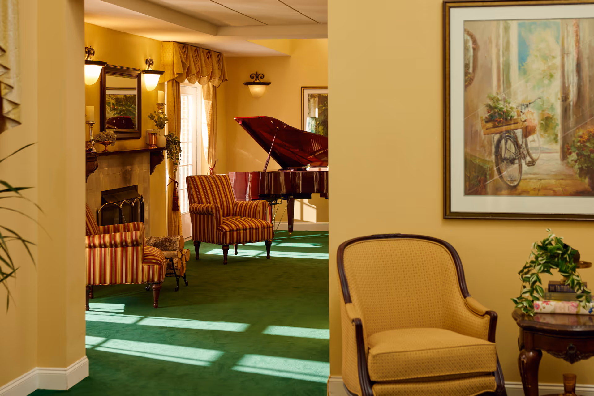 A cozy living room area with yellow walls and green carpet. The room features a striped armchair, a yellow upholstered chair, a wooden side table with books and a plant, a fireplace with decorative items on the mantel, and a grand piano in the background near a window with curtains.