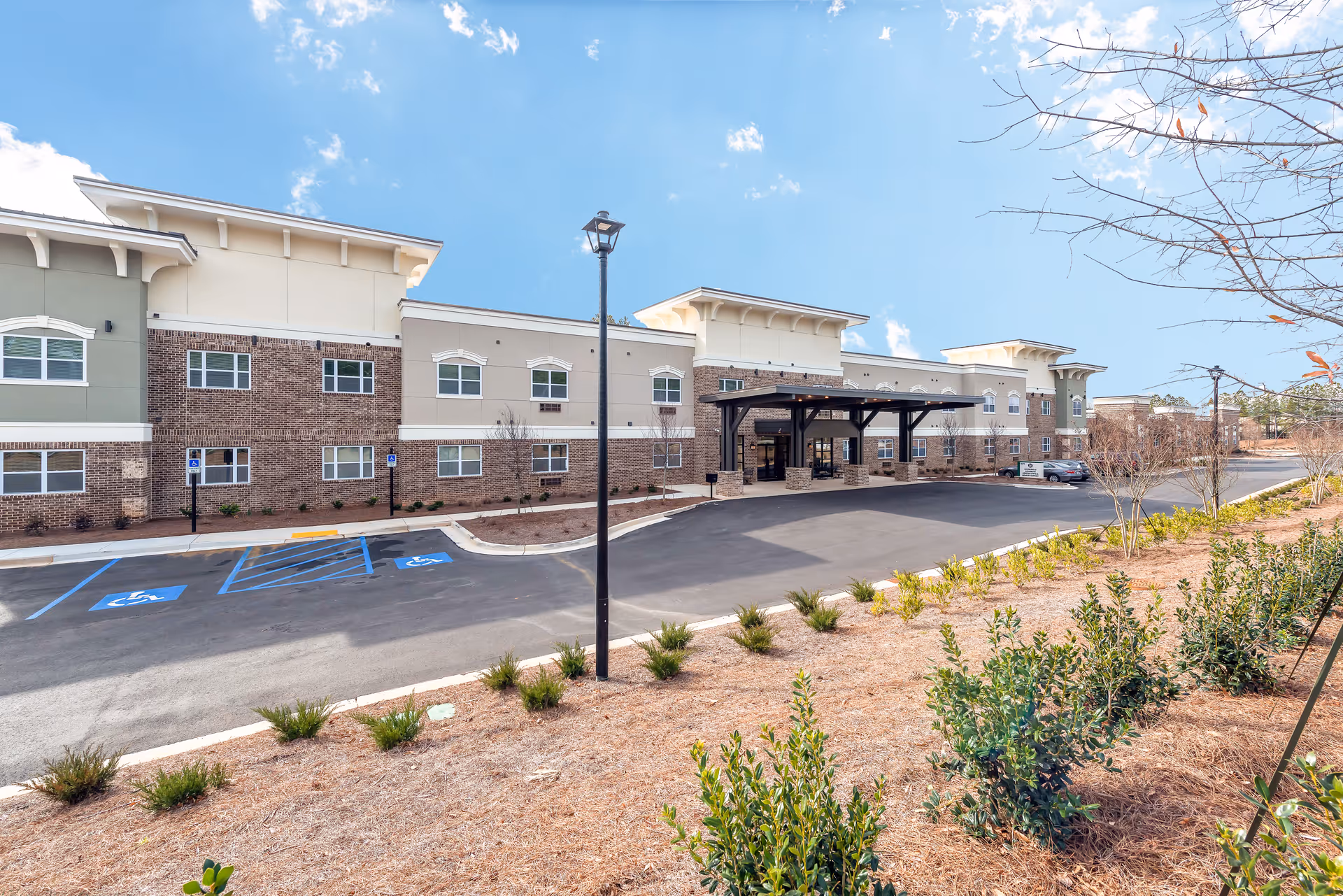 Exterior view of The Addison of Alpharetta senior living facility showing a large two-story building with a covered entrance, parking spaces including handicapped spots, landscaped areas with small bushes and trees, under a partly cloudy blue sky.