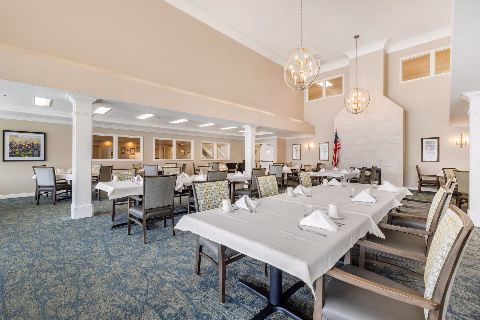 A spacious dining room with multiple tables covered in white tablecloths, each set with napkins, cups, and glasses. The room features high ceilings, modern spherical chandeliers, a large fireplace with an American flag beside it, and patterned carpet flooring. Several chairs with patterned upholstery surround the tables, and framed artwork decorates the walls.
