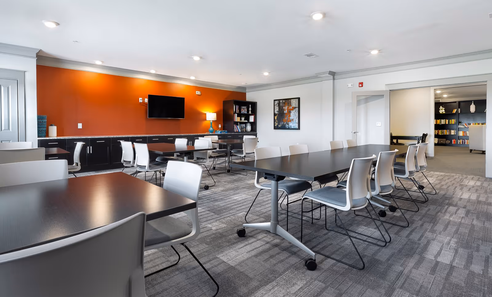 Bright communal activity room with multiple tables and chairs, a wall-mounted TV on an orange accent wall, and shelving in the background.