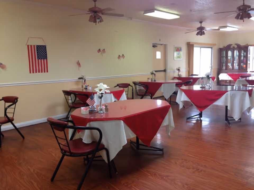 A dining room with several tables covered in white tablecloths and red overlays, each table decorated with small flower arrangements and American flags. The room has wooden flooring, ceiling fans, and an American flag hanging on the wall. There is a china cabinet and a door leading outside or to another room.