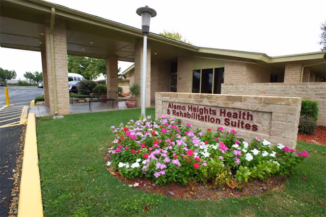 Entrance area of Alamo Heights Health & Rehabilitation Suites with a covered drop-off area, a flower bed with pink, white, and red flowers, and a stone sign displaying the facility name.
