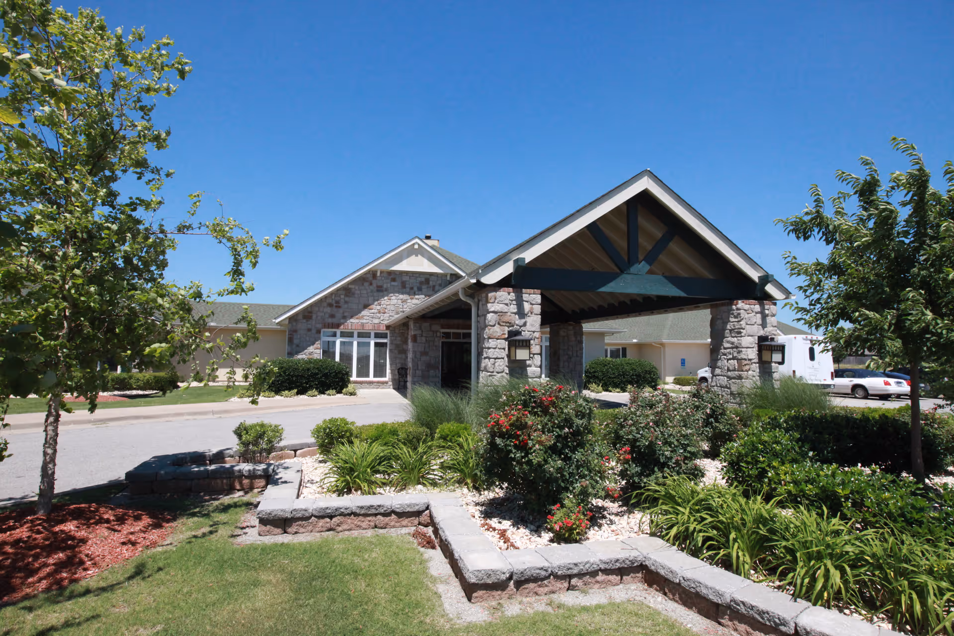 Front entrance of a single-story assisted living facility with a stone portico, landscaped garden beds, and a clear blue sky.