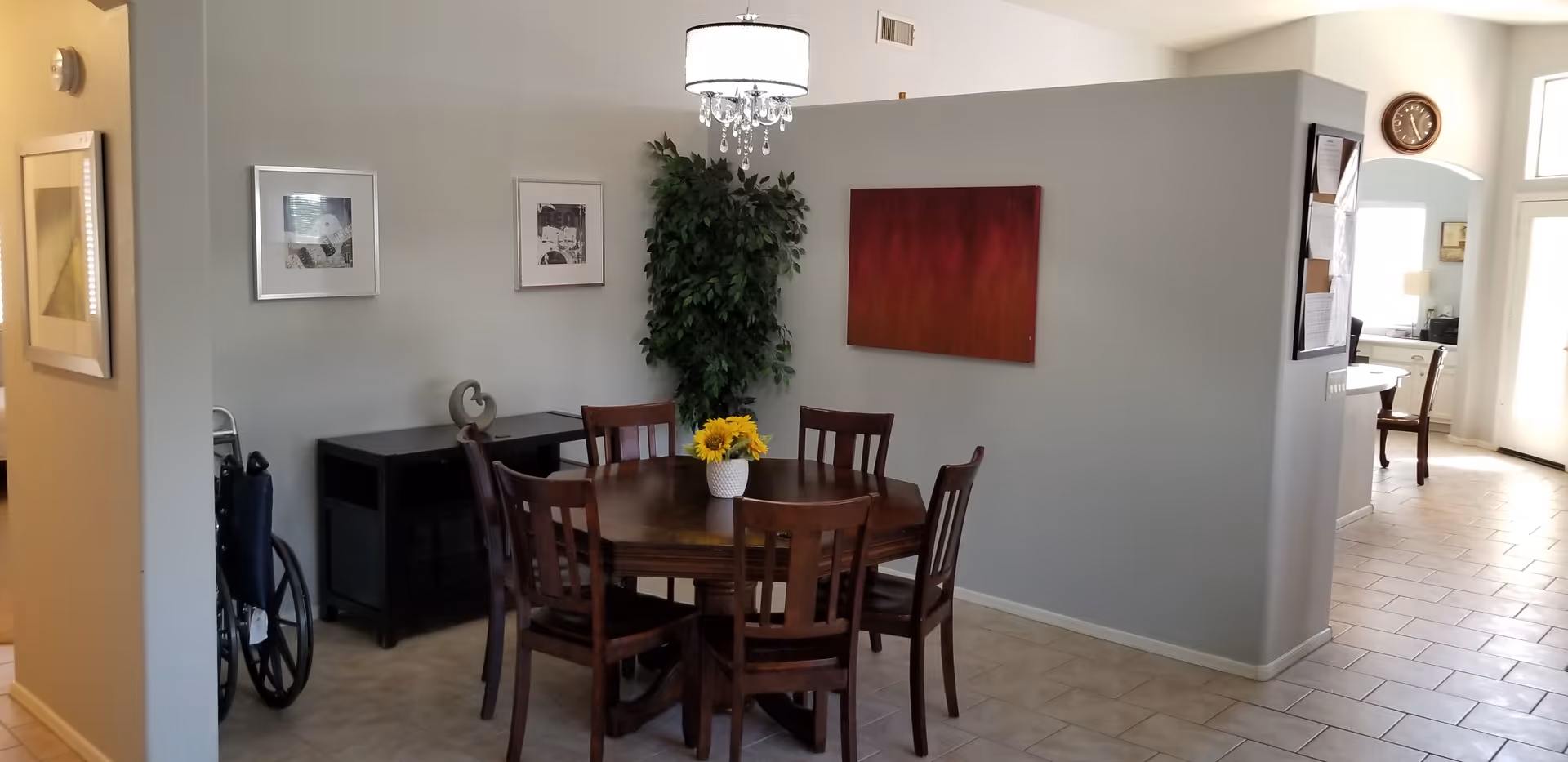 A dining area with a round wooden table surrounded by six wooden chairs. A vase with yellow flowers is placed in the center of the table. The walls are painted light gray and decorated with framed pictures and a red abstract painting. A tall green plant is positioned in the corner. The floor is tiled, and a wheelchair is partially visible to the left. In the background, there is a partial view of a kitchen and a clock on the wall.