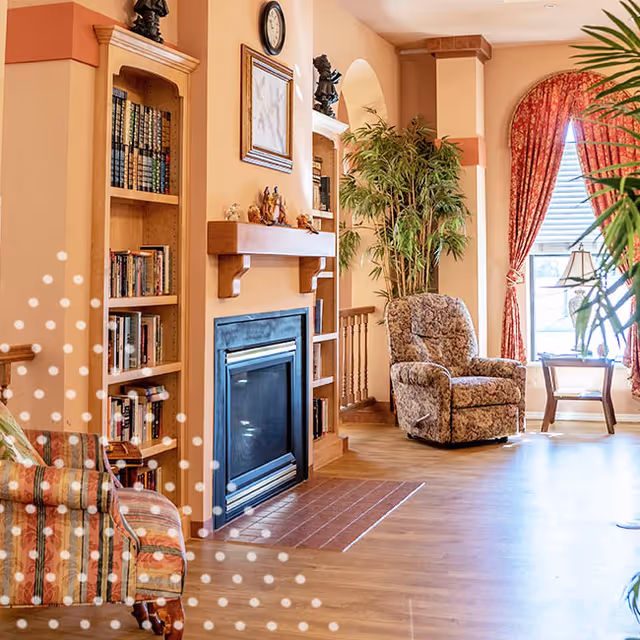 A cozy living room area in an assisted living facility featuring a fireplace with a wooden mantel, built-in bookshelves filled with books, a patterned armchair, a recliner chair, a large potted plant, and a window with red patterned curtains letting in natural light.