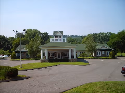 Front exterior of a single-story senior living facility with a covered entrance supported by columns, driveway, and surrounding landscaping.