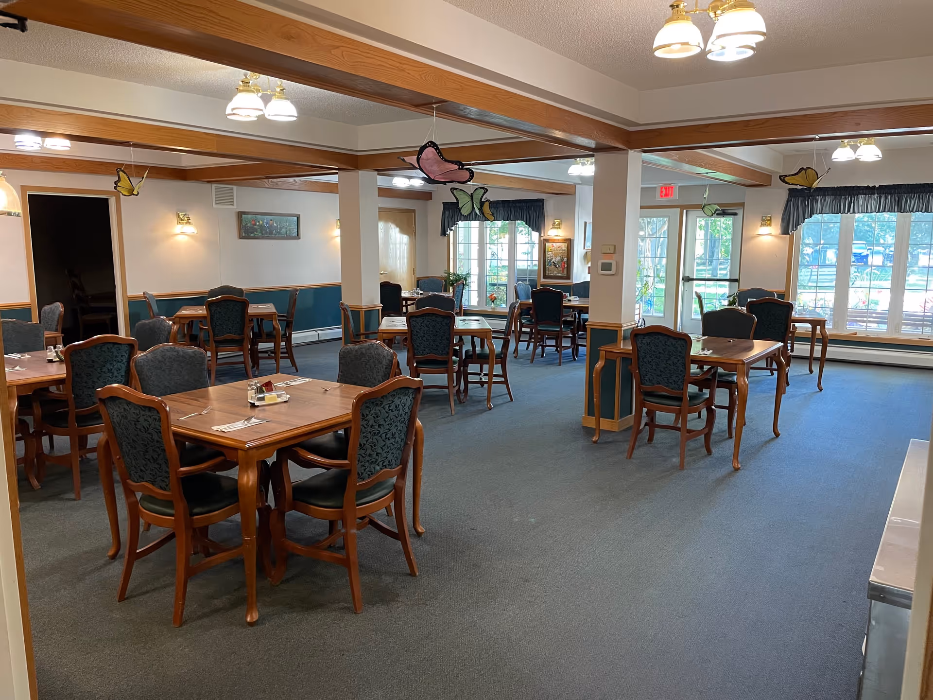 Senior living dining room with several wooden tables and upholstered chairs, windows along the far wall, and hanging light fixtures.