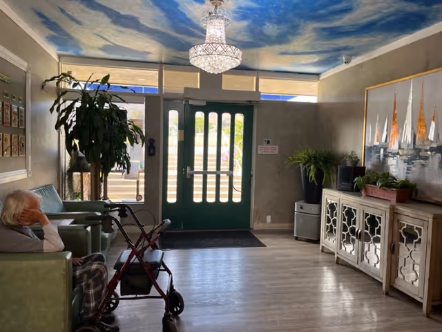 Assisted living lobby with green double entrance doors, seating where a resident sits beside a walker, a chandelier and painted sky ceiling, plants and a decorative console.