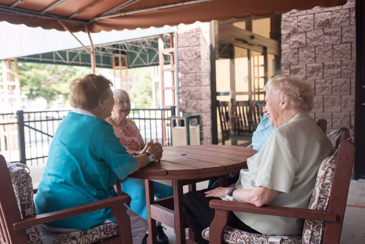 Four elderly women sitting around a wooden outdoor table under a canopy, engaged in conversation and smiling. The setting appears to be a patio area with a stone wall and glass doors in the background.