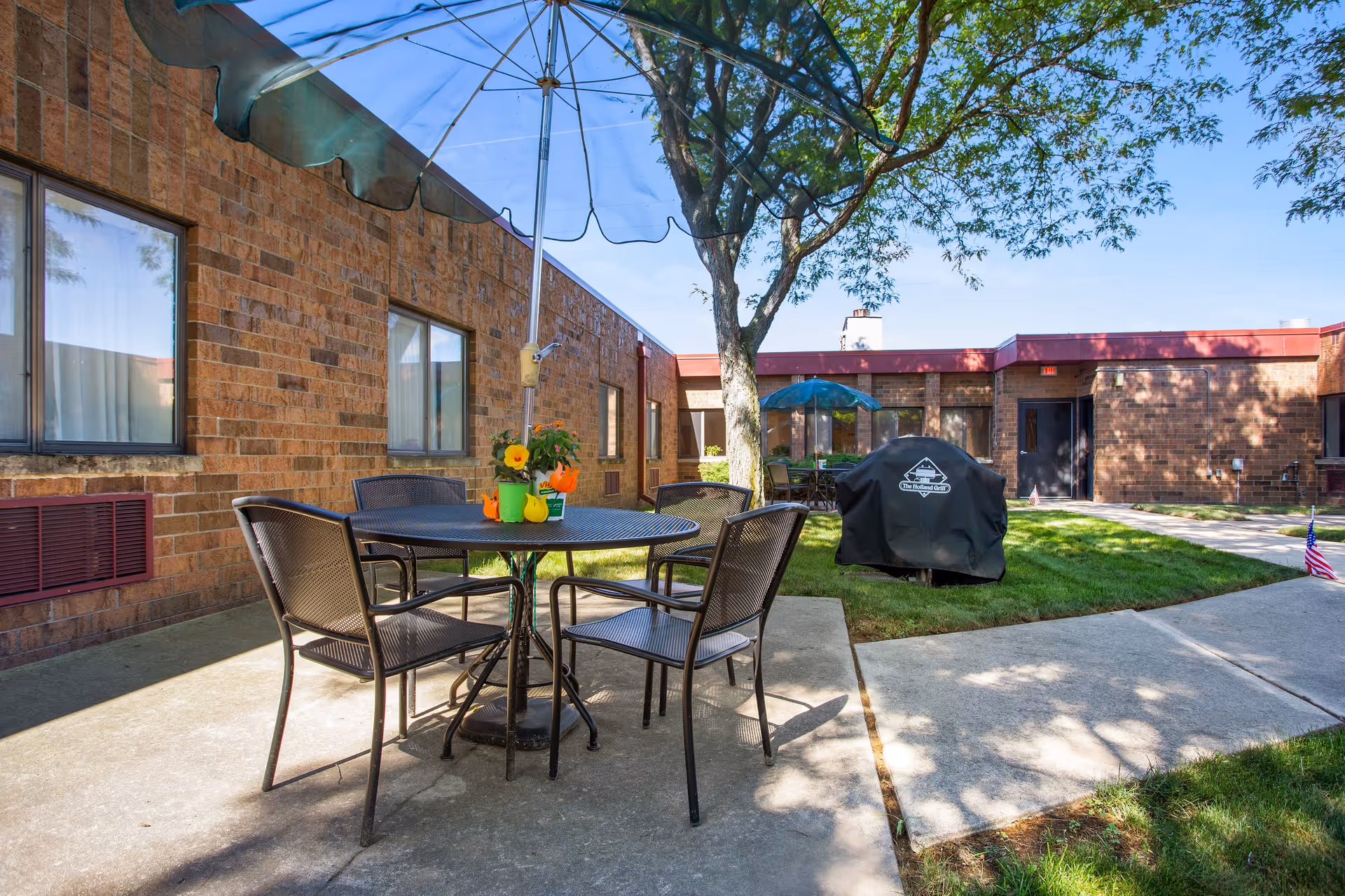 Outdoor patio area at Arbor Inn with a round metal table and four chairs under a large blue umbrella. There are flowers in small pots on the table. The patio is surrounded by a brick building with several windows and a tree providing shade. A covered grill and another table with an umbrella are visible in the background.