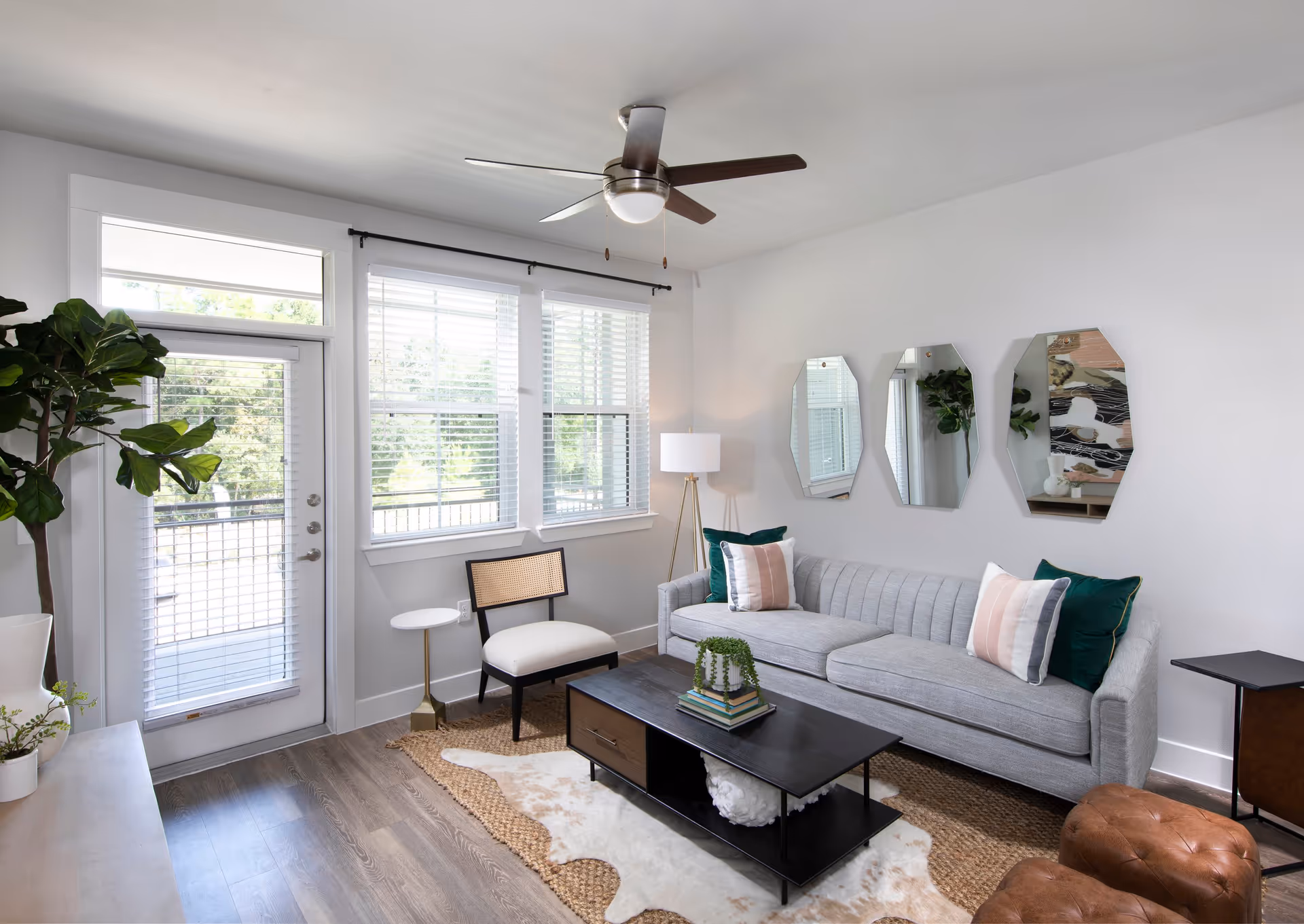 A bright living room with a gray sofa adorned with green and pink pillows, a black coffee table with books and a plant, a white cushioned chair, a floor lamp, three decorative mirrors on the wall, a ceiling fan, and a door with windows letting in natural light.