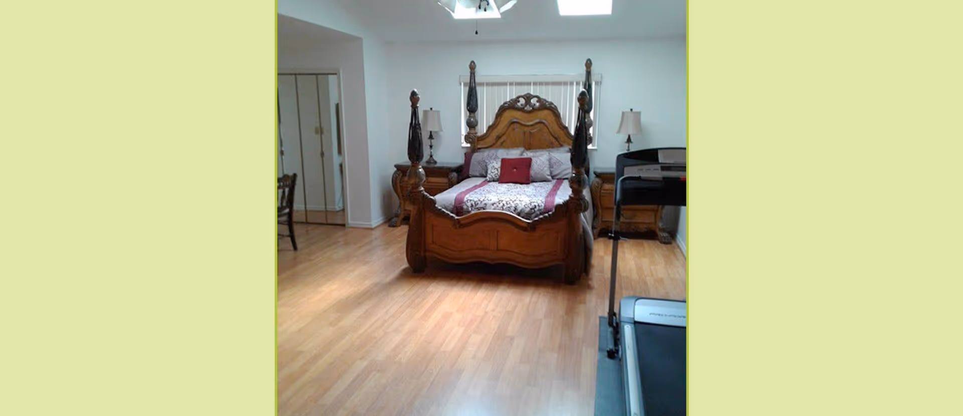A bedroom with a wooden four-poster bed adorned with patterned bedding and pillows. There are two matching wooden nightstands with lamps on either side of the bed. A treadmill is positioned on the right side of the room. The floor is light wood, and there is a mirrored closet door on the left wall. The walls are painted white, and a ceiling fan is visible above.
