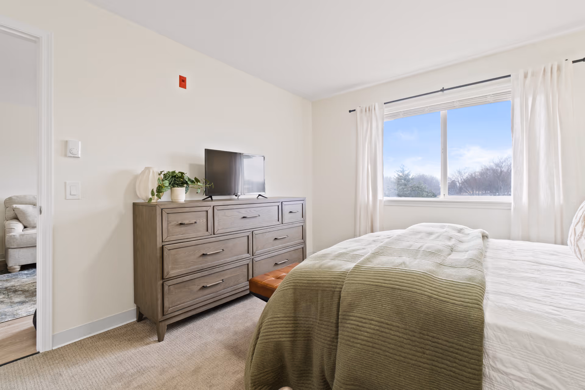 A bright bedroom with a large window covered by white curtains letting in natural light. The room features a bed with a green quilted blanket, a wooden dresser with multiple drawers, a small potted plant, and a flat-screen TV on top. The walls are painted white, and a glimpse of a living room with a beige sofa is visible through an open door.