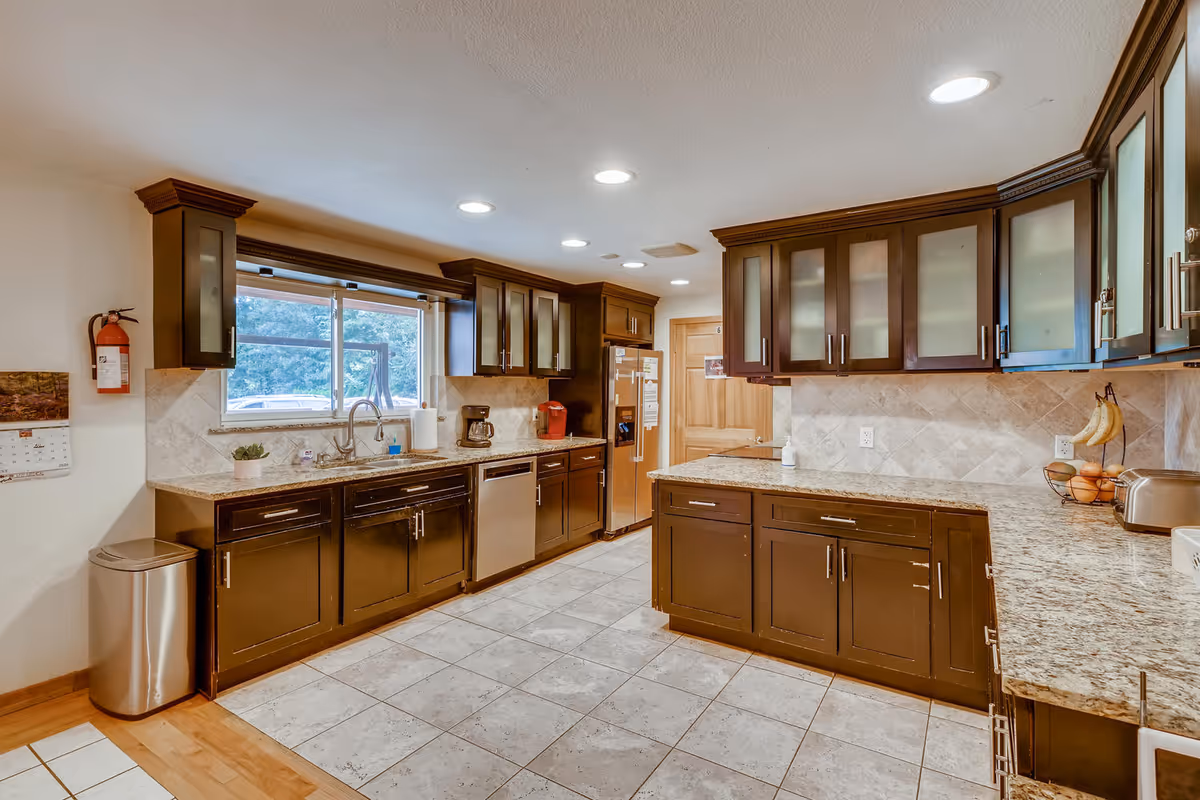 A spacious kitchen with dark wood cabinets and granite countertops. The kitchen features a large window above the sink, stainless steel dishwasher, refrigerator, coffee maker, and a Keurig machine. The floor is tiled, and the ceiling has recessed lighting. There is a fire extinguisher mounted on the wall and a trash can near the entrance.