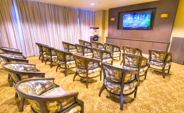 A small theater or media room with three rows of patterned armchairs facing a wall-mounted flat screen TV. The room has beige carpeting, a popcorn machine in the corner, and floor-to-ceiling curtains covering the windows.