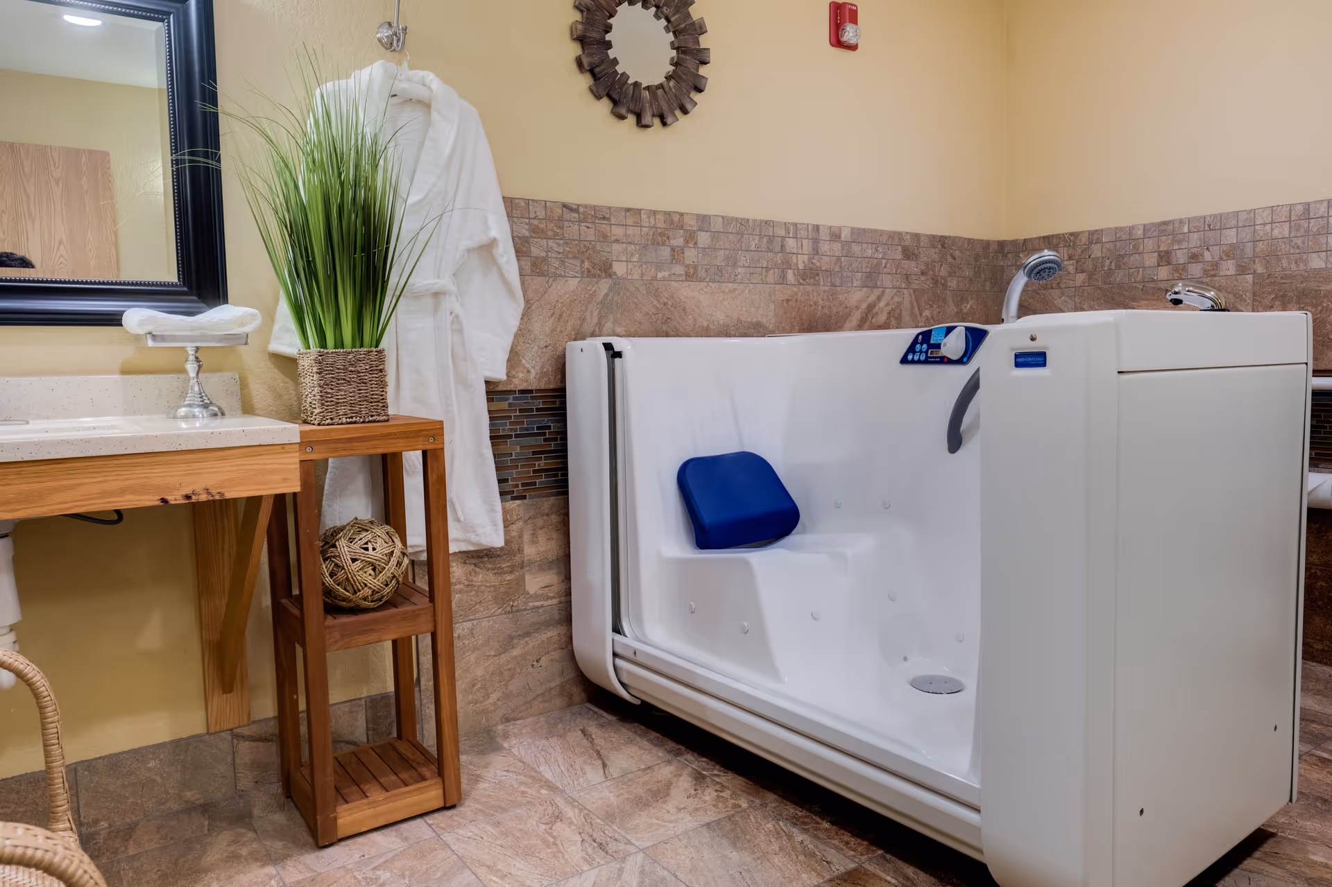 A bathroom with a walk-in bathtub featuring a blue seat cushion and control panel. Next to the bathtub is a wooden shelf holding a decorative plant and a woven ball. A white bathrobe hangs on the wall above the shelf. The walls are tiled halfway up with beige and brown tiles, and there is a mirror and countertop with a towel on the left side.