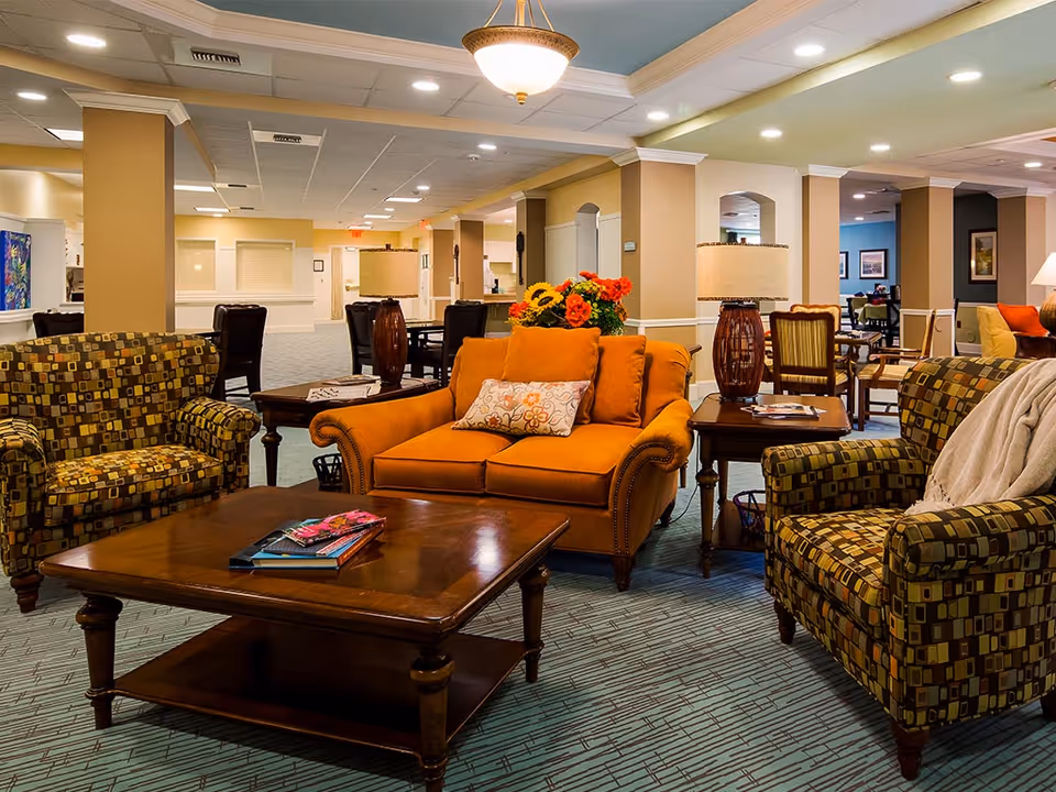 A spacious, well-lit senior living lounge with patterned armchairs, an orange loveseat, wooden coffee tables, and decorative lamps.