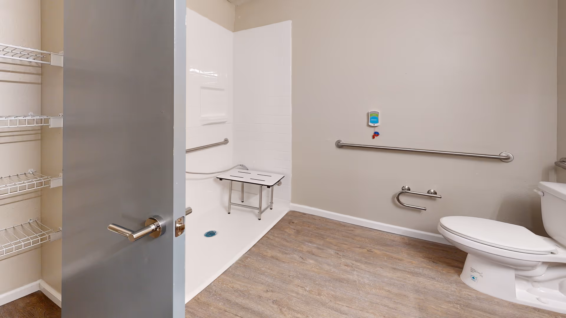 Accessible bathroom with a walk-in shower featuring a white foldable shower bench and grab bars. The bathroom has a toilet with grab bars on the wall beside it, wood-look flooring, and a partially open gray door with wire shelving visible on the left.