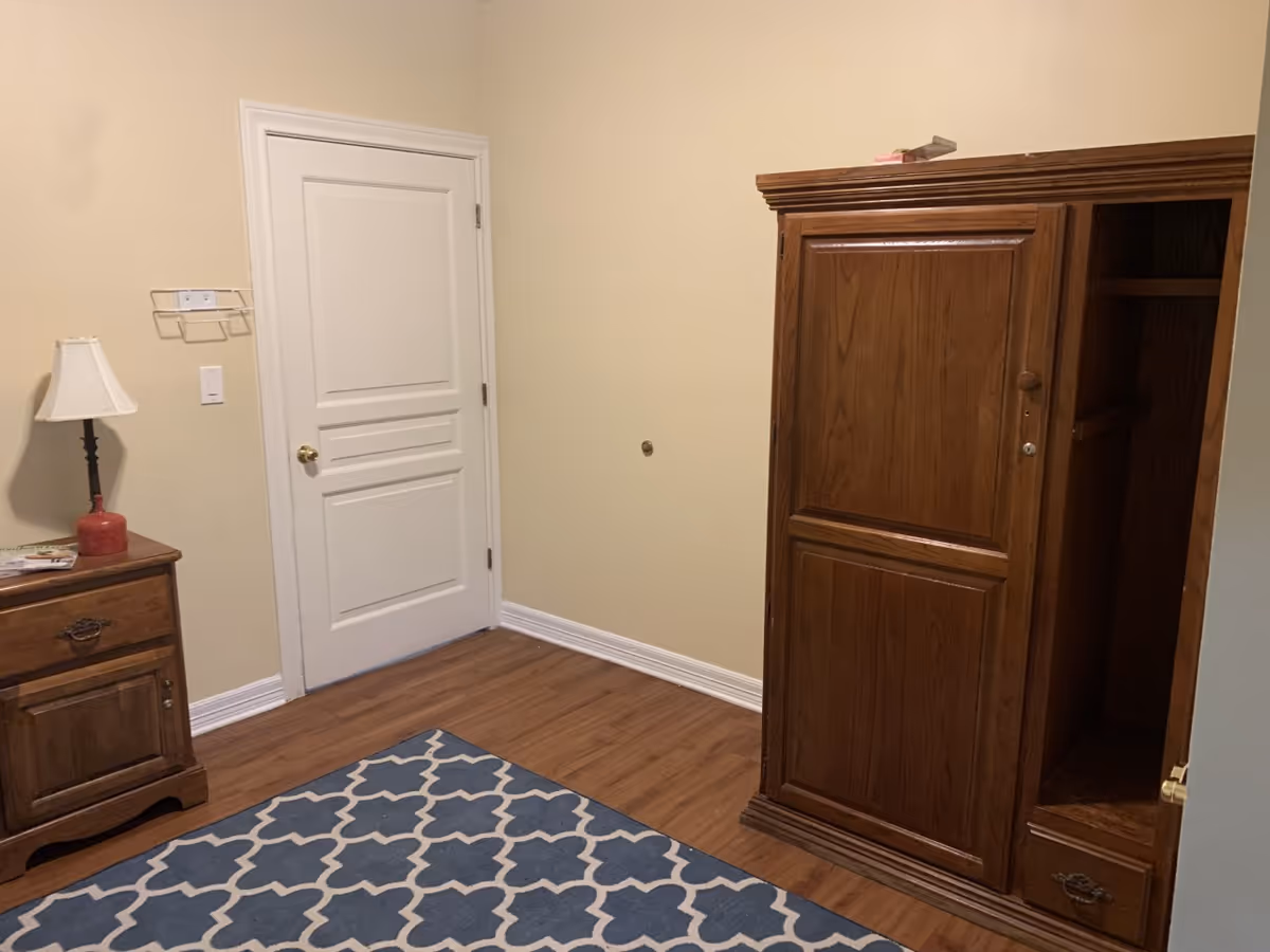 A beige room features a wooden wardrobe, nightstand, lamp, and a blue geometric rug.
