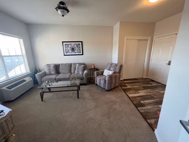 A living room area with a gray sofa adorned with patterned pillows, a matching patterned armchair, and a glass coffee table with decorative items on it. There is a framed artwork on the wall above the sofa, a window with blinds on the left side, and a small round side table between the sofa and armchair. The floor is carpeted except for the entryway area, which has wood-style flooring. The walls are painted white.