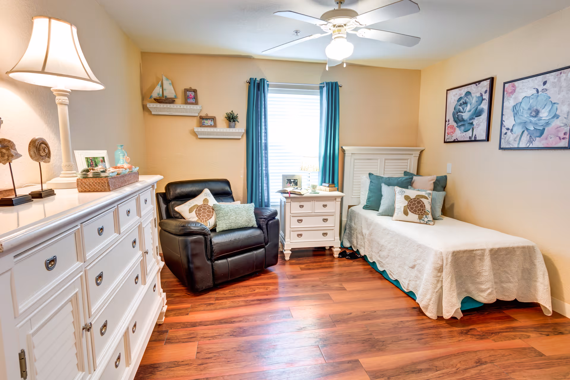 A cozy bedroom with a single bed covered in a white quilt and multiple decorative pillows, including one with a turtle design. Next to the bed is a white nightstand with books and a lamp. A black leather armchair with pillows is positioned near the window, which has teal curtains. A white dresser with decorative items and a lamp is on the left side of the room. The walls are painted beige and adorned with two framed floral paintings. The floor is wooden, and a ceiling fan with a light is mounted on the ceiling.